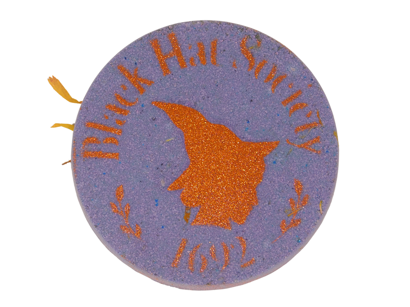circular purple bath bomb with an autumn orange glitter airbrush of a witch's side profile surrounded by text that says black hat society 1692